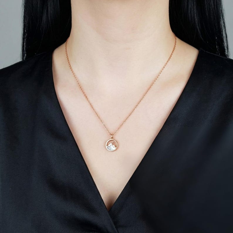 Puede incluir: Un collar de oro rosa con un colgante circular con una escena de monta&ntilde;a. El colgante tiene un dise&ntilde;o de monta&ntilde;a blanco y plateado dentro de un marco de oro rosa. El collar se usa sobre una blusa negra.