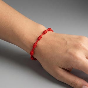 May include: A red knotted bracelet is worn on a wrist. The bracelet is made of a woven material and features several evenly spaced knots. The background is a solid gray.