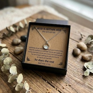 Puede incluir: Un collar plateado con un colgante de monta&ntilde;a, presentado en una caja de regalo negra. La caja contiene una tarjeta con el texto "GOD IS WITHIN HER, She will not fall" y texto inspirador. El collar se muestra sobre una superficie de madera.