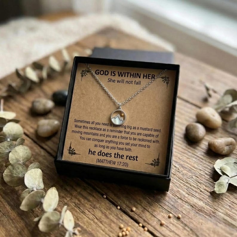 Puede incluir: Un collar plateado con un colgante de monta&ntilde;a, presentado en una caja de regalo negra. La caja contiene una tarjeta con el texto "GOD IS WITHIN HER, She will not fall" y texto inspirador. El collar se muestra sobre una superficie de madera.
