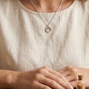 Puede incluir: Un collar de plata con un colgante redondo con un peque&ntilde;o dise&ntilde;o. El collar cuelga sobre una blusa color crema. Una peque&ntilde;a botella de vidrio con un tap&oacute;n de corcho es visible en primer plano.