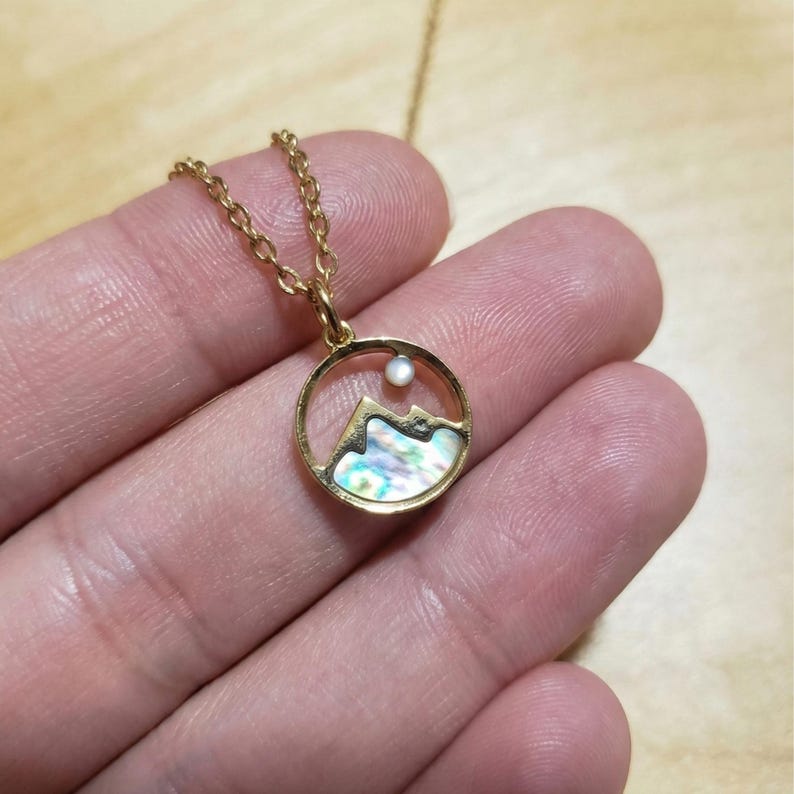 Puede incluir: Collar dorado con un colgante circular con la silueta de una monta&ntilde;a y una incrustaci&oacute;n iridiscente. Una peque&ntilde;a perla redonda est&aacute; engastada encima de la monta&ntilde;a. El collar tiene una cadena delicada.