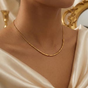 May include: A delicate gold necklace with a series of small, cylindrical and spherical beads. The necklace is worn on a person's neck, against a backdrop of a cream-colored fabric. The necklace has a simple, elegant design.