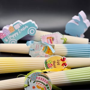 May include: Easter-themed decorative candles with colorful tags and ribbons. The candles are white with pastel color accents, and the tags feature Easter bunny and egg designs. A light blue truck and bunny-shaped cutouts are also present.