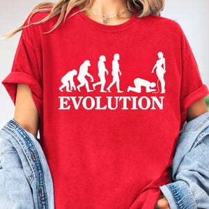 May include: Red t-shirt with a white graphic depicting the evolution of humans, ending with a person kneeling. The word "EVOLUTION" is printed in white below the graphic. The shirt has rolled-up sleeves.