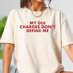 May include: A cream-colored t-shirt with the red text "MY DUI CHARGES DON'T DEFINE ME." The shirt has short sleeves and a relaxed fit. The person is wearing a gold necklace and bracelets.