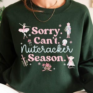 May include: Dark green sweatshirt with the text "Sorry. Can't. Nutcracker Season." in pink and white. The design includes illustrations of a ballerina, nutcracker, mouse, and snowflakes. A festive holiday-themed top.