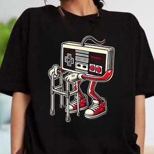 May include: Black t-shirt featuring a cartoon illustration of a retro video game controller with arms, legs, and red sneakers, using a walker. The controller has a D-pad, buttons, and the word "Nintendo" on it.