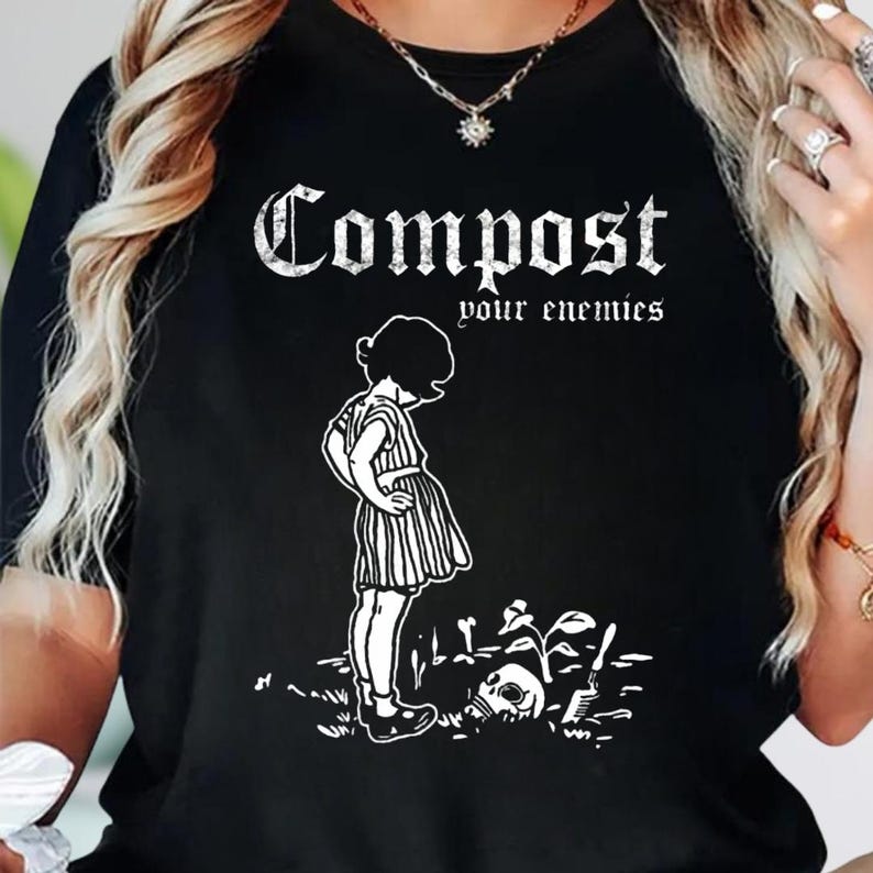 May include: A black t-shirt featuring a white graphic print of a person standing over a skull and a small plant. The text "Compost your enemies" is printed in a distressed white font above the illustration, which also includes a small shovel.