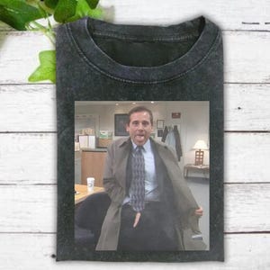 May include: Dark gray t-shirt featuring a framed image of a man in a suit and tie, with his mouth open and a surprised expression. The man is wearing a long coat and is seated in an office setting.