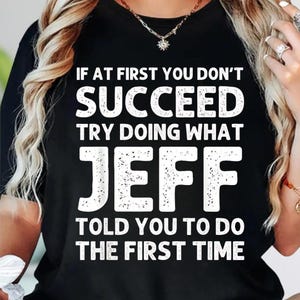 May include: Black t-shirt with white text that reads: "IF AT FIRST YOU DON'T SUCCEED TRY DOING WHAT JEFF TOLD YOU TO DO THE FIRST TIME." The shirt has a crew neck and is likely made of cotton.
