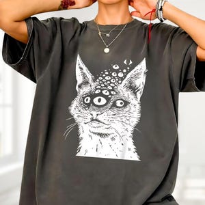 以下が含まれることがあります： 複数の目を持つ猫の白いイラストが特徴の濃い灰色のTシャツ。猫のグラフィックは詳細で風変わりなデザインです。Tシャツは半袖で、カジュアルウェアに適したリラックスしたフィット感です。
