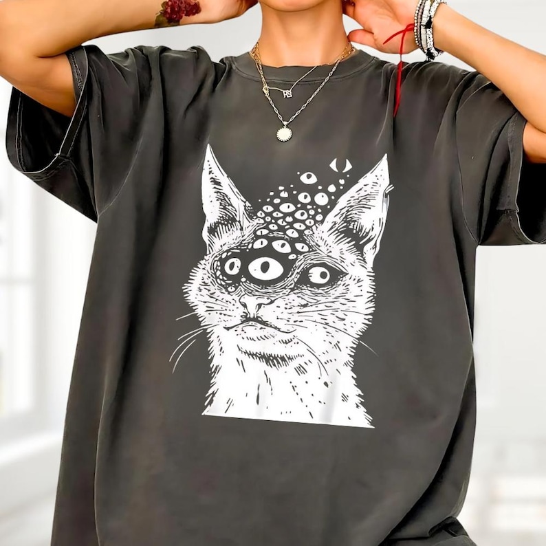 以下が含まれることがあります： 複数の目を持つ猫の白いイラストが特徴の濃い灰色のTシャツ。猫のグラフィックは詳細で風変わりなデザインです。Tシャツは半袖で、カジュアルウェアに適したリラックスしたフィット感です。