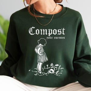 May include: A dark forest green crewneck sweatshirt featuring a white graphic print. The design displays the text "Compost your enemies" in a gothic font above an illustration of a child standing near a skull and a small plant in the dirt.