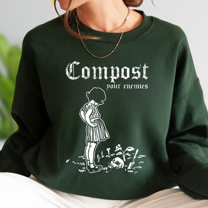 May include: A dark forest green crewneck sweatshirt featuring a white graphic print. The design displays the text "Compost your enemies" in a gothic font above an illustration of a child standing near a skull and a small plant in the dirt.