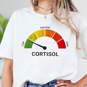 May include: White t-shirt with a graphic of a cortisol level meter. The meter has a black needle pointing to the green "low" section. The text "CORTISOL" is printed below the meter in black.