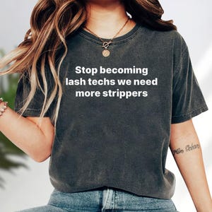 May include: A charcoal grey short-sleeve t-shirt featuring the white printed text Stop becoming lash techs we need more strippers. The garment has a crew neckline and a vintage-style washed finish, paired with blue denim jeans and a gold necklace.