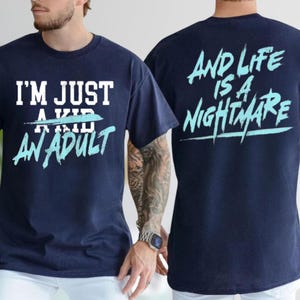 A man wearing a navy blue t-shirt with the text "I'M JUST A KID AN ADULT" printed on it, along with a tattoo on his arm.