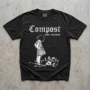 May include: A black short-sleeved t-shirt featuring a white graphic print of a child standing in a garden next to a skull and a small plant. The text "Compost your enemies" is printed in a white gothic-style font above the illustration.