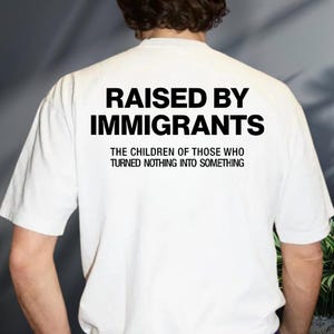 May include: White t-shirt with the text "RAISED BY IMMIGRANTS" in bold black letters. Below the main text is the phrase "THE CHILDREN OF THOSE WHO TURNED NOTHING INTO SOMETHING" in smaller black letters.