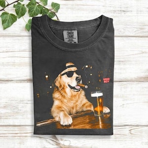 May include: A charcoal gray t-shirt featuring a graphic of a golden retriever wearing black sunglasses and a baseball cap, holding a cigar in its mouth while resting its paws on a wooden bar next to a glass of beer with the text GOOD BOY.