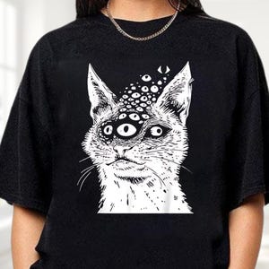 以下が含まれることがあります： 黒いクルーネックのトレーナー。白いイラストは、複数の目を持つ猫が描かれています。猫の顔にはヒゲと少し開いた口が描かれています。デザインはシャツの前面の中央に配置されています。