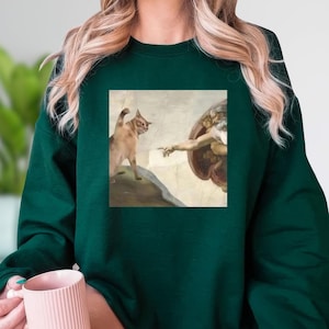 A woman wearing a green sweater with a printed image of a cat and a human figure, possibly depicting the biblical creation of Adam.