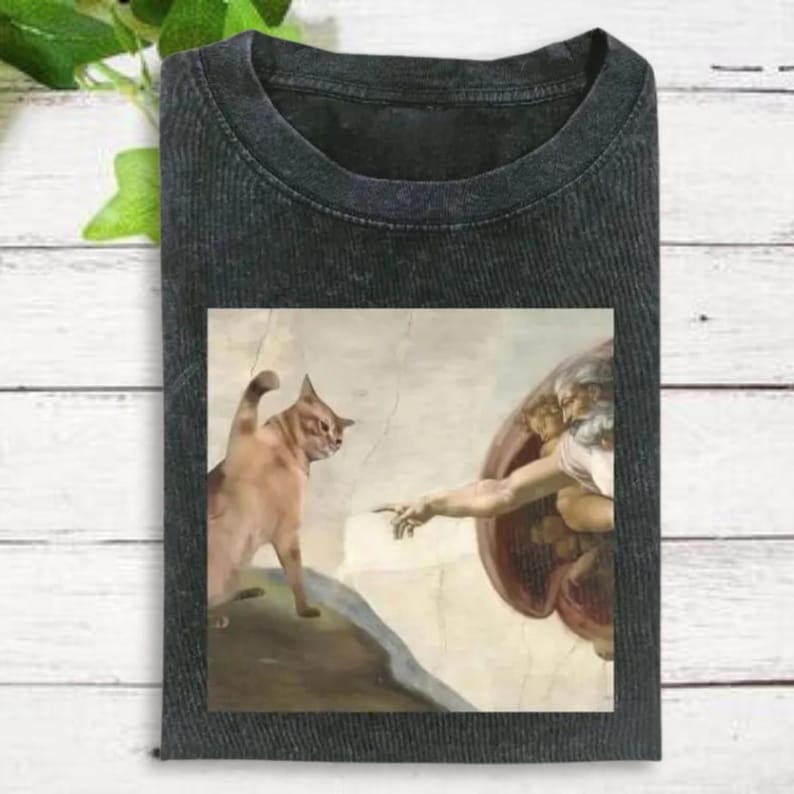 a black t-shirt with a printed design depicting a painting of a cat and a human figure, possibly referencing the biblical creation of Adam.