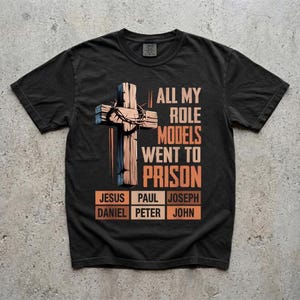 May include: A black short-sleeve t-shirt featuring a graphic of a wooden cross with a crown of thorns. The text reads: ALL MY ROLE MODELS WENT TO PRISON, followed by a grid of names: JESUS, PAUL, JOSEPH, DANIEL, PETER, and JOHN in orange boxes.