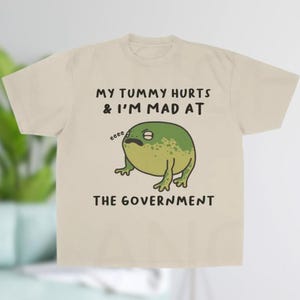 K&ouml;nnte beinhalten: Beiges T-Shirt mit dem Text "MY TUMMY HURTS & I'M MAD AT THE GOVERNMENT" und einem Cartoon-Frosch. Der Frosch ist gr&uuml;n mit dunkleren gr&uuml;nen Flecken und hat einen traurigen Gesichtsausdruck.