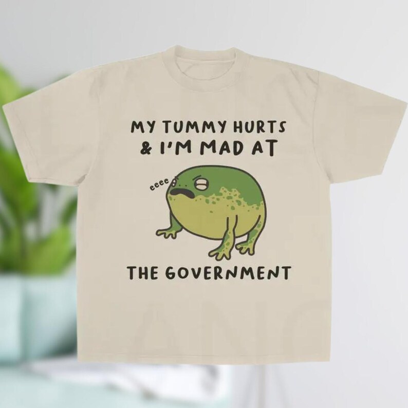 K&ouml;nnte beinhalten: Beiges T-Shirt mit dem Text "MY TUMMY HURTS & I'M MAD AT THE GOVERNMENT" und einem Cartoon-Frosch. Der Frosch ist gr&uuml;n mit dunkleren gr&uuml;nen Flecken und hat einen traurigen Gesichtsausdruck.