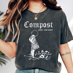 May include: A charcoal grey short-sleeve t-shirt featuring a white graphic print of a person standing next to a skull and a small plant. The text "Compost your enemies" is printed in a white gothic-style font above the illustration.