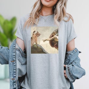 A woman wearing a gray t-shirt with a printed image of a cat and a human figure, possibly depicting the biblical creation of Adam.