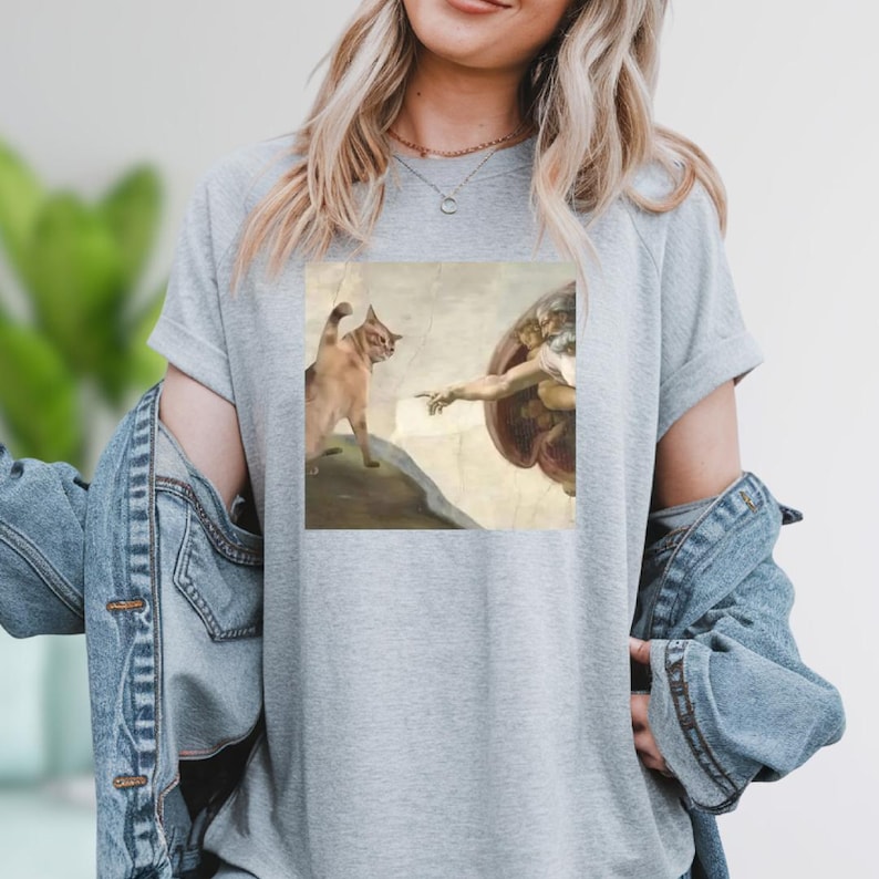 A woman wearing a gray t-shirt with a printed image of a cat and a human figure, possibly depicting the biblical creation of Adam.