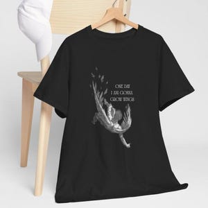 Może przedstawiać: Czarny t-shirt z grafiką spadającego anioła i białym napisem "ONE DAY I AM GONNA GROW WINGS". Koszulka wisi na drewnianym wieszaku.