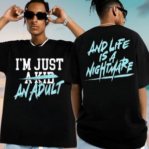 A man wearing a black t-shirt with the text "I'M JUST A KID AN ADULT" and the phrase "AND LIFE IS A NIGHTMARE" printed on it.