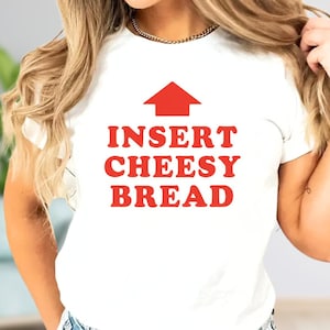 May include: White t-shirt with the text "INSERT CHEESY BREAD" in red, with a red arrow pointing upwards. The shirt is a casual style, suitable for everyday wear.
