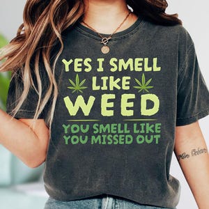 May include: Dark gray t-shirt with the text "YES I SMELL LIKE WEED" in lime green, along with cannabis leaf graphics. The bottom of the shirt reads "YOU SMELL LIKE YOU MISSED OUT".