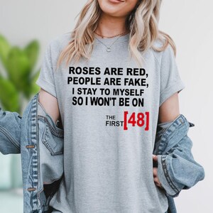 A woman wearing a gray t-shirt with text that says "Roses are red, people are fake, I stay to myself so I won't be on THE FIRST 481".