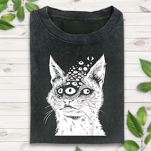 以下が含まれることがあります： 複数の目を持つ猫の白いイラストが描かれた、濃い灰色のTシャツ。猫の顔にはひげと少し不気味な表情が描かれています。Tシャツは、緑の葉のある明るい木製の表面に折りたたまれています。