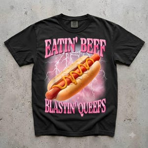 May include: A black short-sleeve t-shirt featuring a graphic of a hot dog with yellow mustard against a pink lightning background. The text EATIN' BEEF is printed in pink above the hot dog, and BLASTIN' QUEEFS is printed in pink below it.