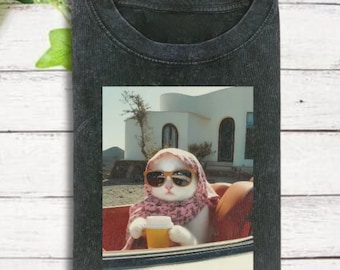 May include: Dark gray t-shirt featuring a graphic of a white cat wearing sunglasses and a scarf, holding a yellow cup. The cat is in a car with a white building in the background. The shirt is made of a soft material.