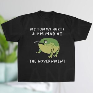 K&ouml;nnte beinhalten: Schwarzes T-Shirt mit einer Cartoon-Frosch-Illustration. Der Frosch ist gr&uuml;n und gelb mit einem m&uuml;rrischen Gesichtsausdruck. Der Text auf dem Shirt lautet "MY TUMMY HURTS & I'M MAD AT THE GOVERNMENT."