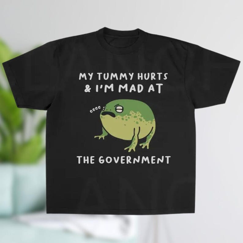 K&ouml;nnte beinhalten: Schwarzes T-Shirt mit einer Cartoon-Frosch-Illustration. Der Frosch ist gr&uuml;n und gelb mit einem m&uuml;rrischen Gesichtsausdruck. Der Text auf dem Shirt lautet "MY TUMMY HURTS & I'M MAD AT THE GOVERNMENT."
