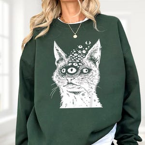以下が含まれることがあります： 複数の目を持つ猫の白いイラストが特徴の濃い緑色のスウェットシャツ。猫のグラフィックは、詳細で少し不気味なデザインです。スウェットシャツは無地で、クルーネックです。