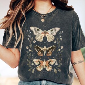 May include: A charcoal gray short-sleeve t-shirt featuring a graphic print of three moths arranged vertically, surrounded by delicate botanical illustrations of leaves and small flowers in shades of cream, brown, and muted green.
