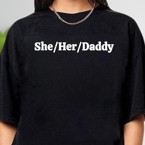 May include: Black crew neck t-shirt with the text "She/Her/Daddy" in white block letters. The shirt is made of a soft material and is a casual style. A silver chain necklace is visible at the neckline.