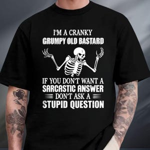May include: A black short-sleeved t-shirt featuring a white graphic of a skeleton with arms raised. The text reads: I'M A CRANKY GRUMPY OLD BASTARD IF YOU DON'T WANT A SARCASTIC ANSWER DON'T ASK A STUPID QUESTION. The design is centered on the chest.