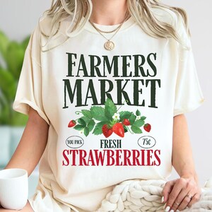 May include: Cream-colored t-shirt with the text "FARMERS MARKET" in dark green. Below the text is a graphic of strawberries and leaves. The words "FRESH STRAWBERRIES" are in red. The shirt has short sleeves.