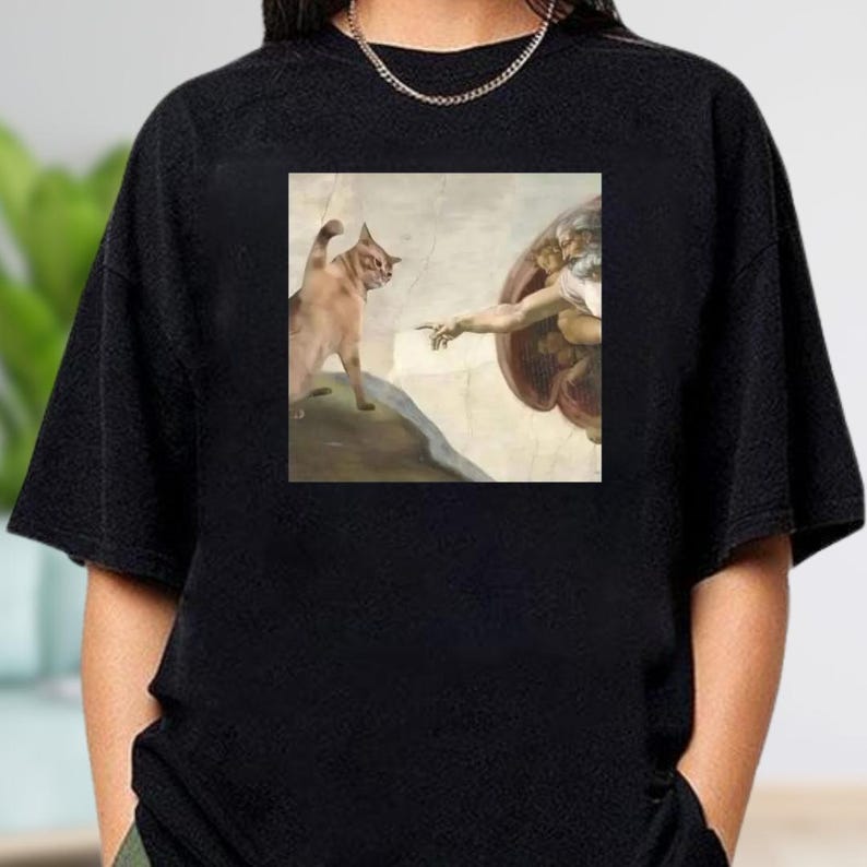 A black t-shirt with a square graphic depicting a cat and a human figure, possibly representing the biblical scene of the creation of Adam.